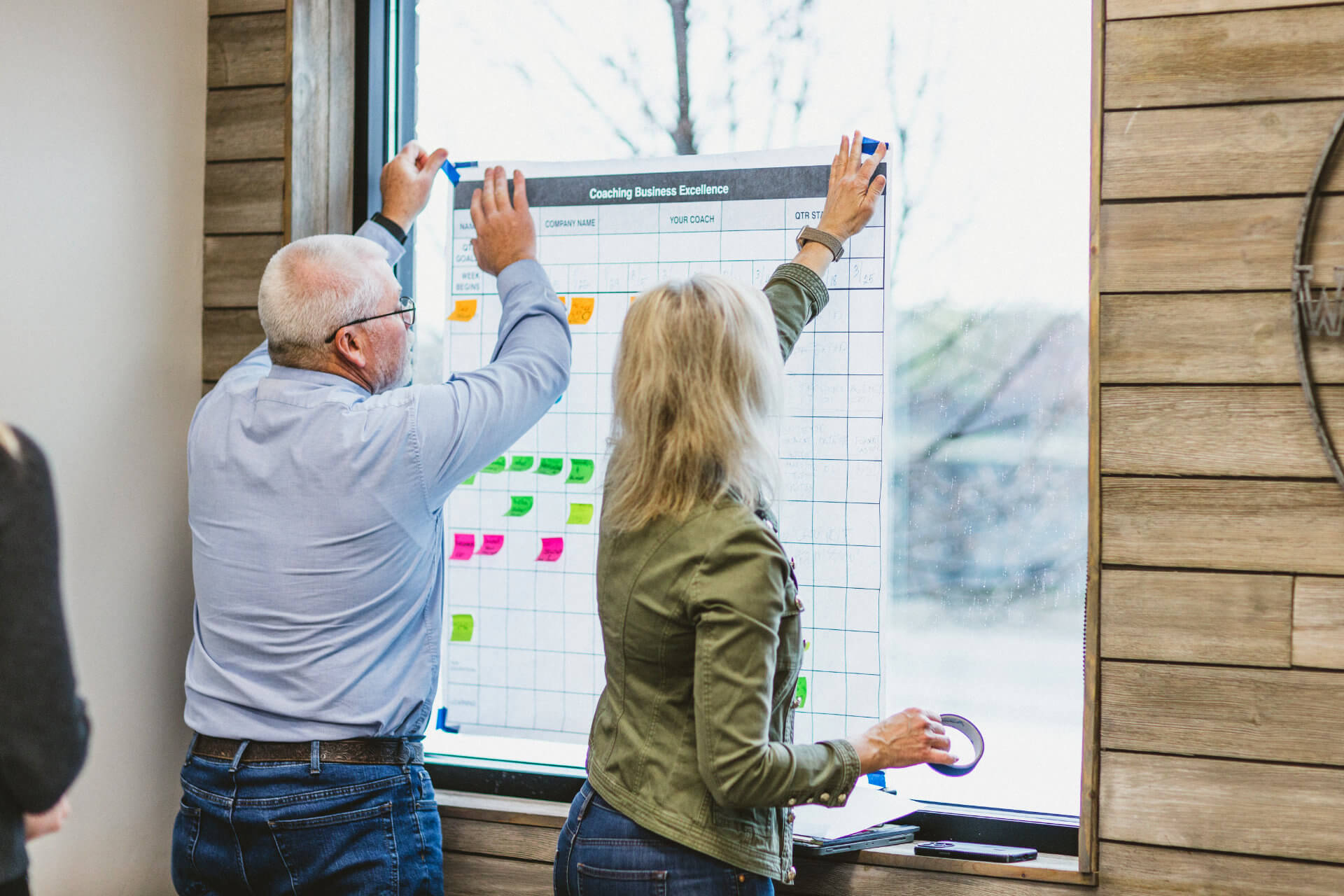 Two people hanging a Coaching Business Excellence planning board covered in sticky notes