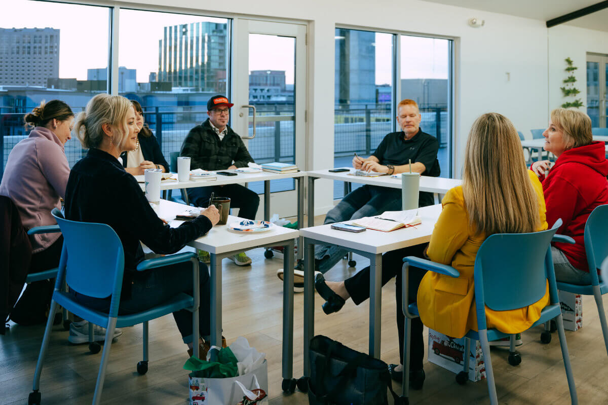 Six professionals seated around tables in a bright conference room with city views