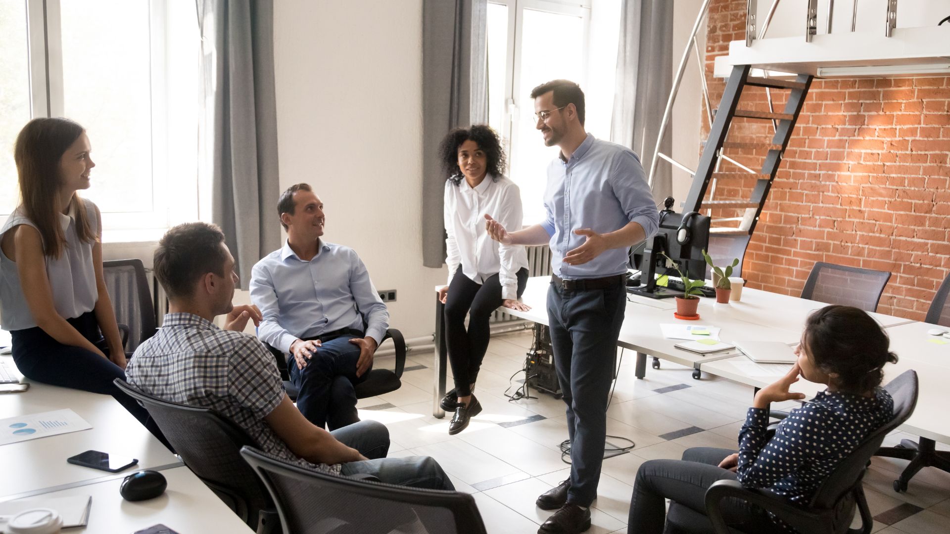 Six professionals in a casual standing meeting in a modern office with an exposed brick wall