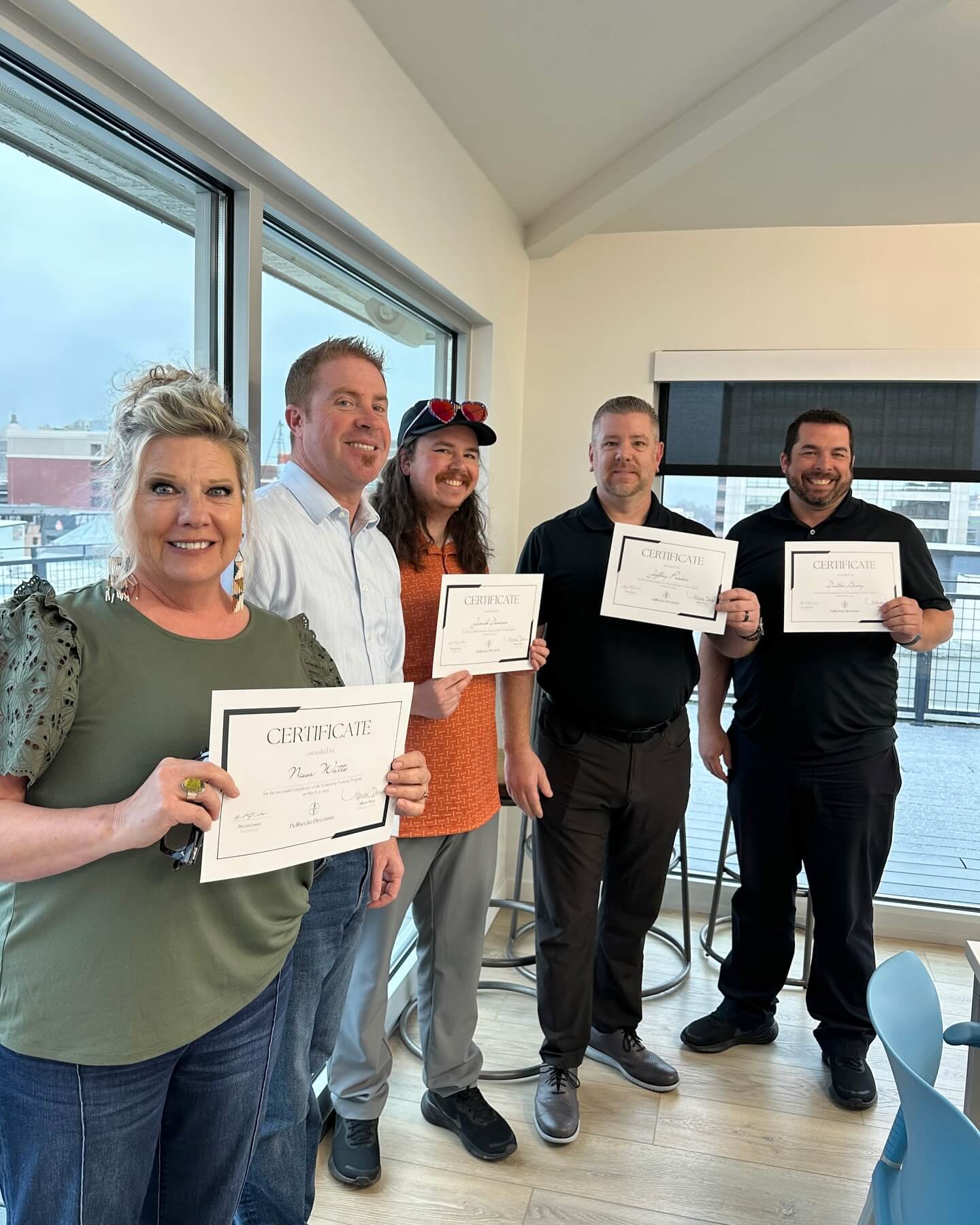 Five participants holding certificates of completion in a bright office with city views