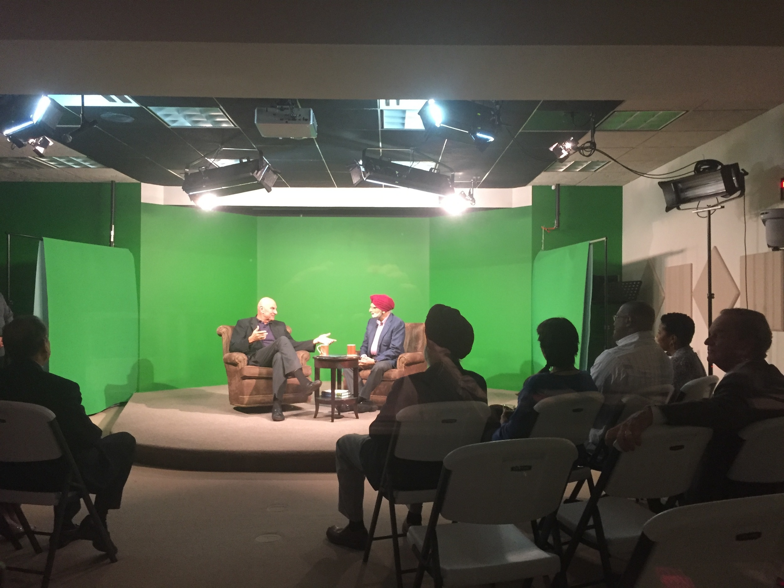 Ratanjit S. Sondhe and a host seated in armchairs on a green-screen television studio set with an audience watching