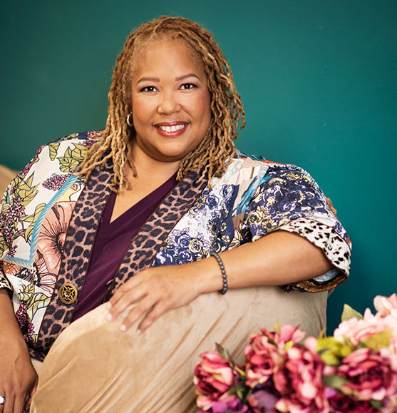 Dr. Réquel Lopes in a floral jacket smiling on a couch beside flowers