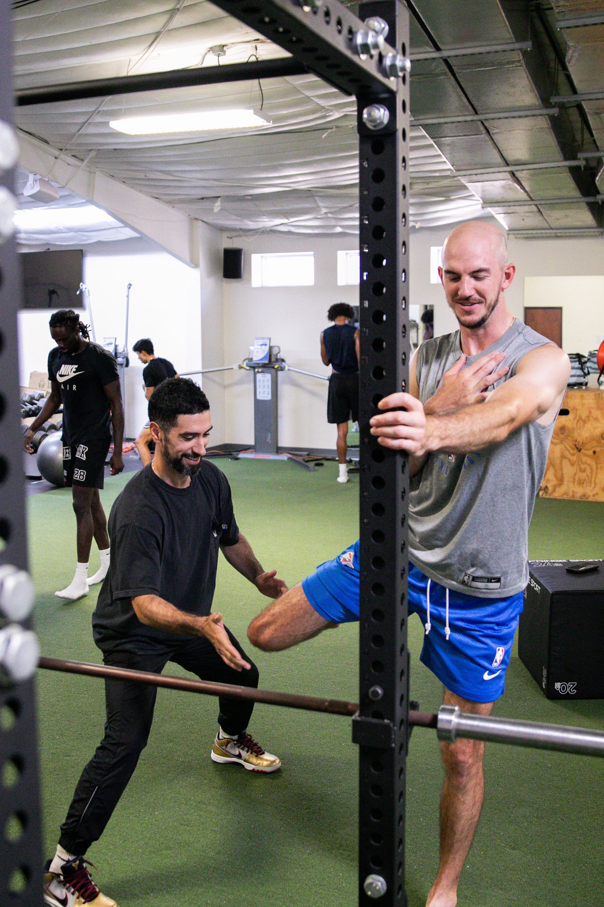 Trainer coaching Alex Caruso through a leg raise at a squat rack with athletes training behind them