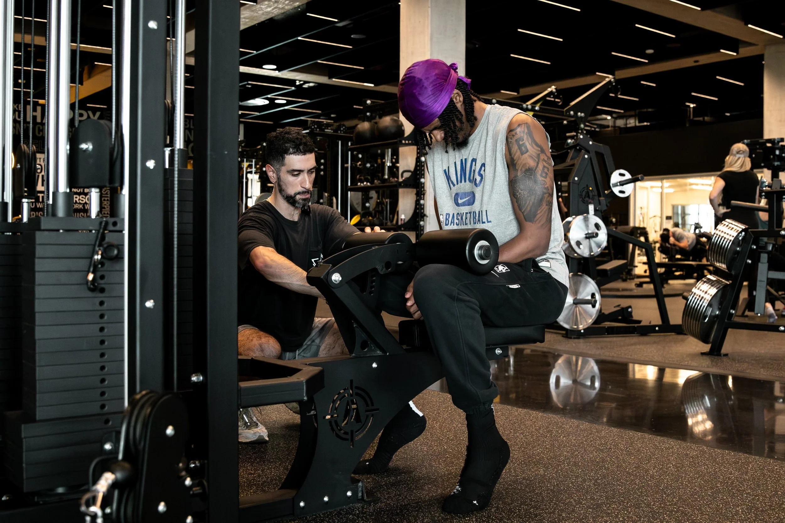 Durable Athlete trainer adjusting a leg-curl machine for an NBA player in Sacramento Kings gear inside a high-end gym