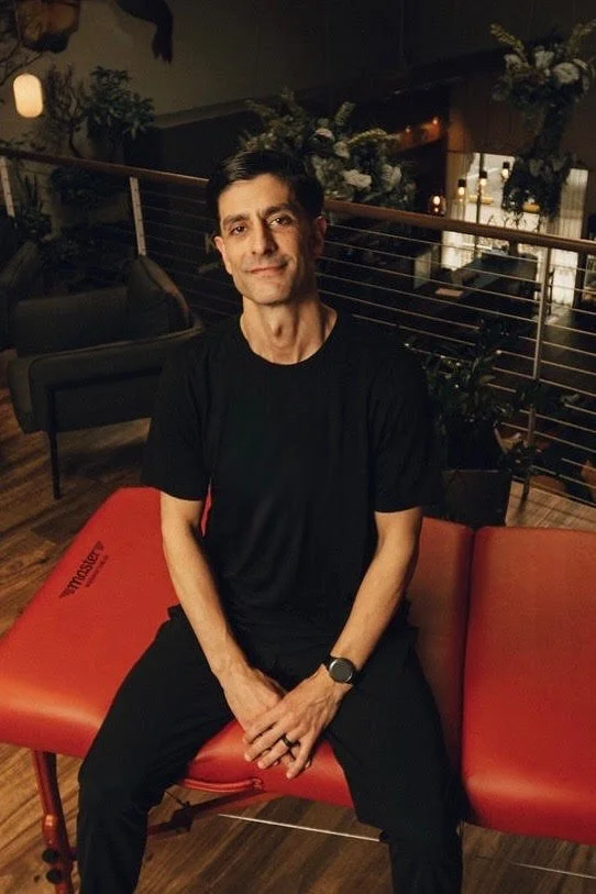 Timothy Lucas seated on a red massage table in a plant-filled lounge