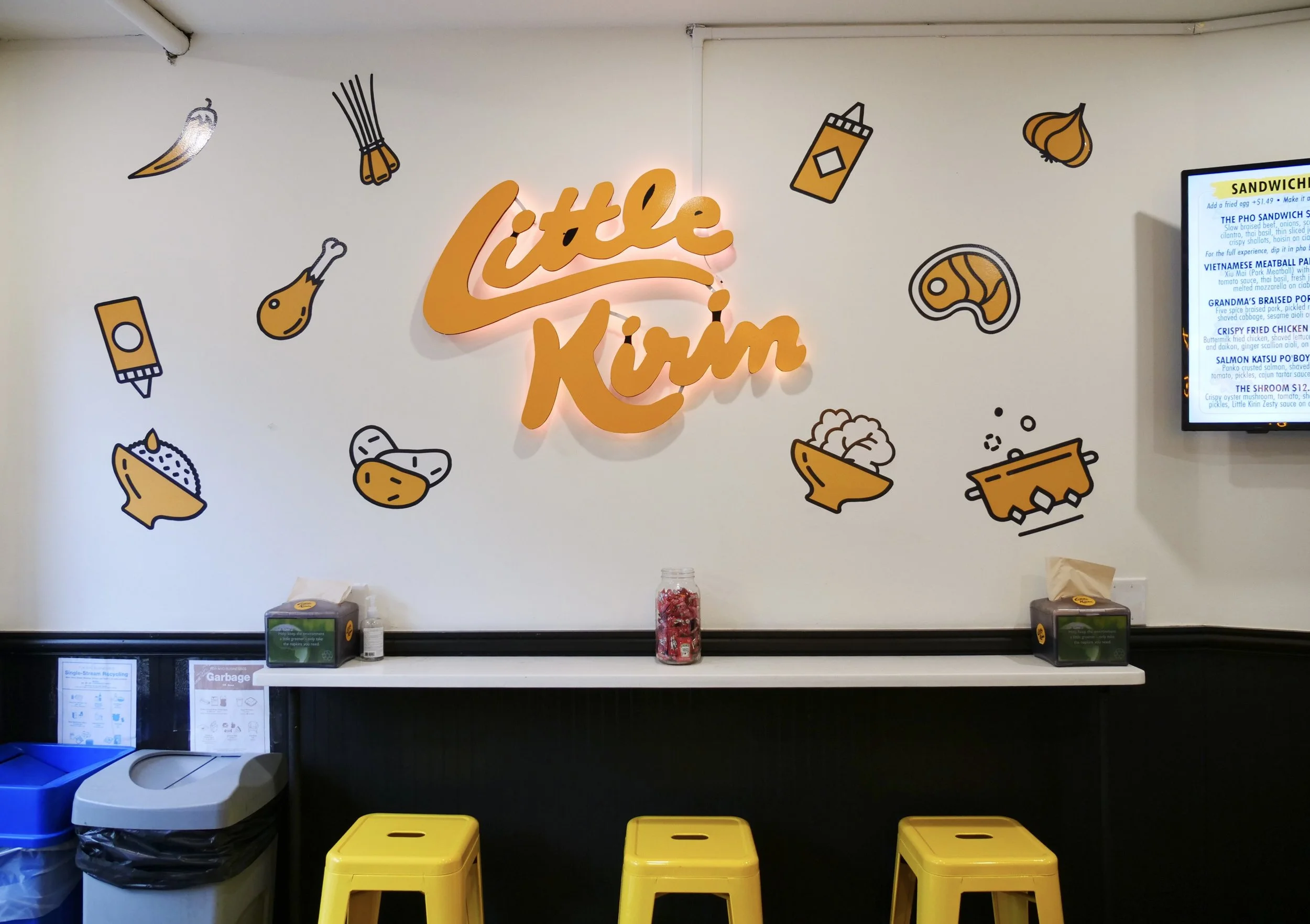 Little Kirin dining room: illuminated wall logo with illustrated food icons and yellow counter stools