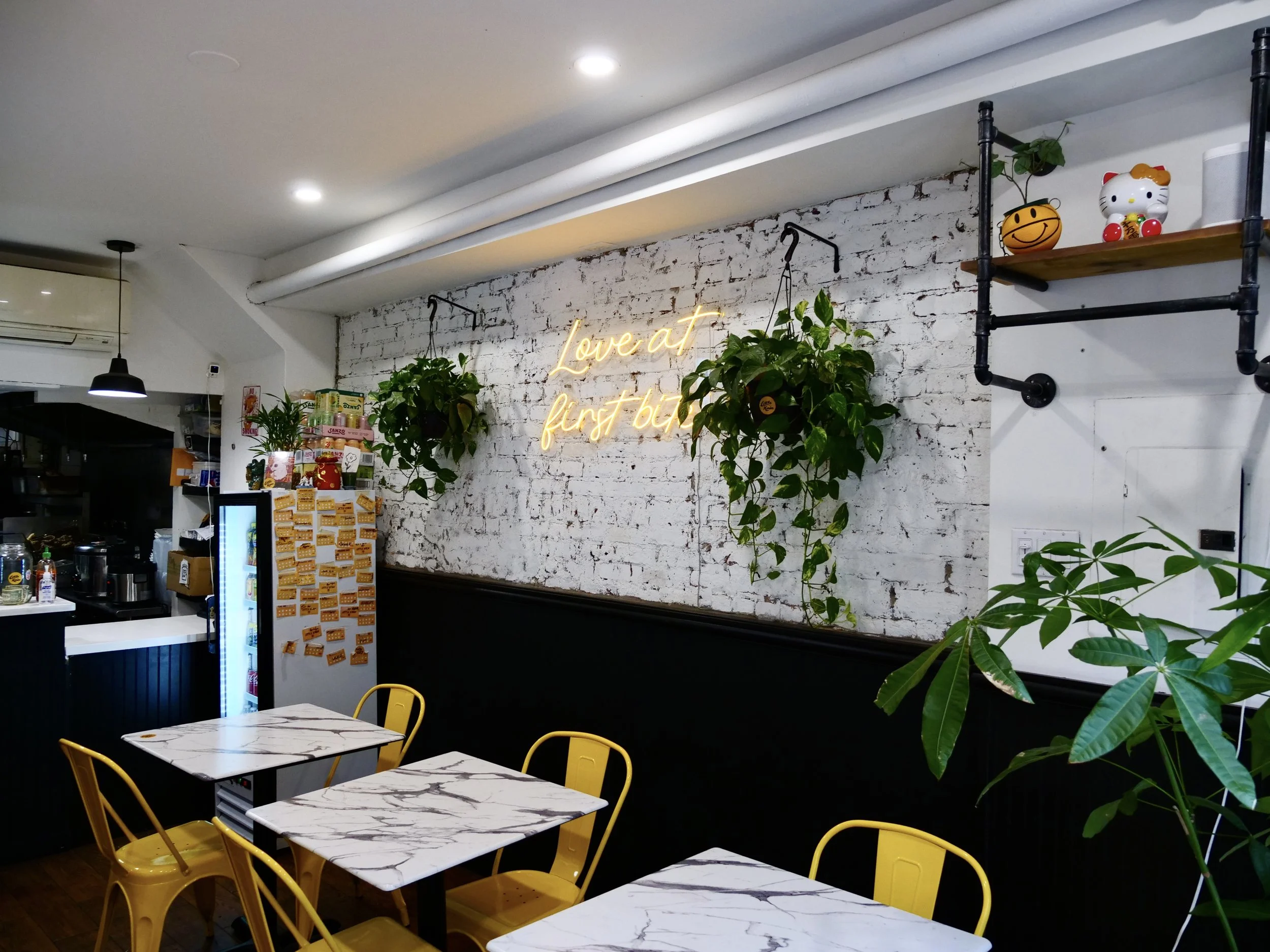 Little Kirin dining room with white painted brick, neon Love at first bite sign, hanging plants, yellow chairs and marble tables