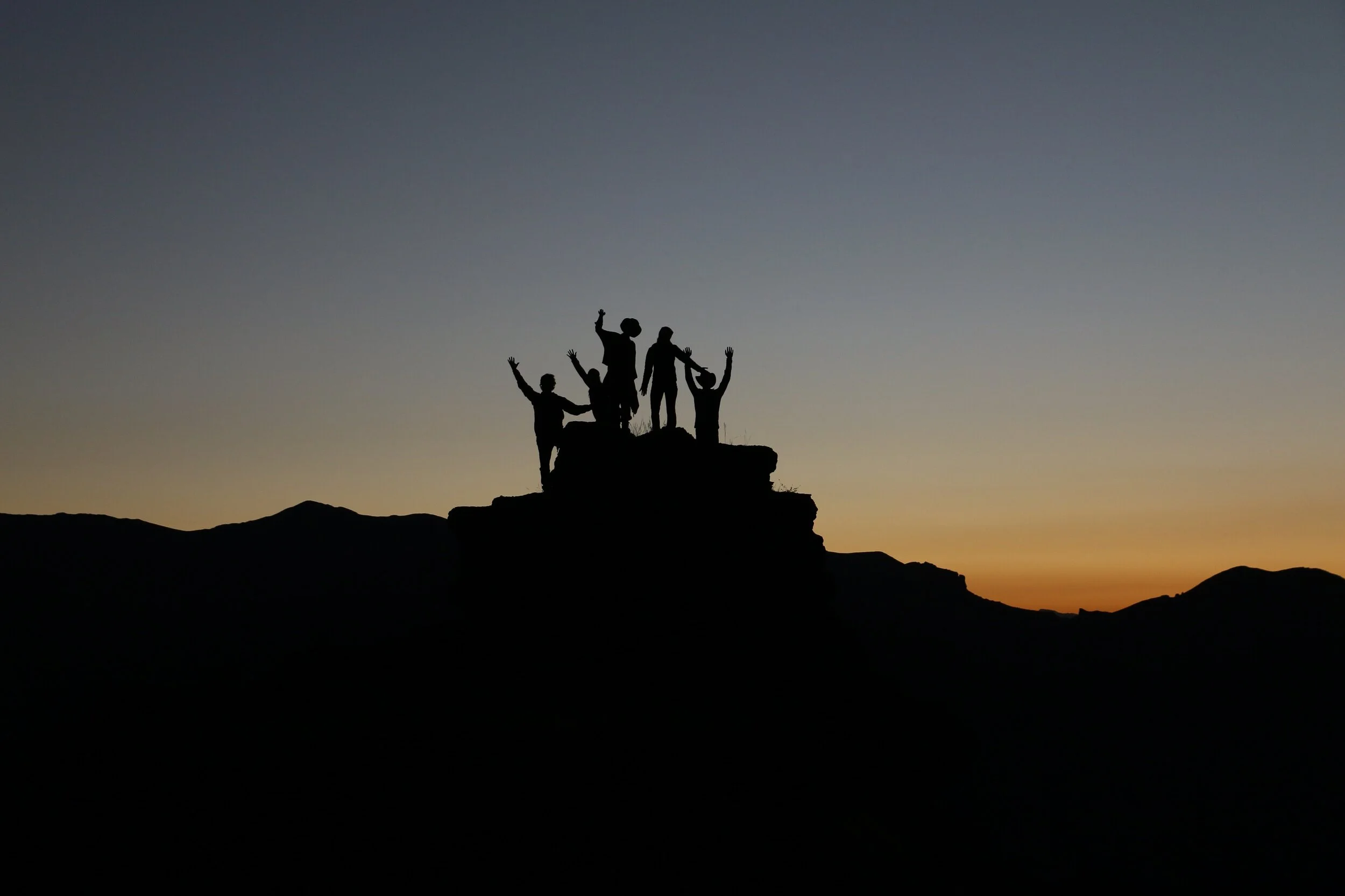 Four people with arms raised on a hilltop at sunset