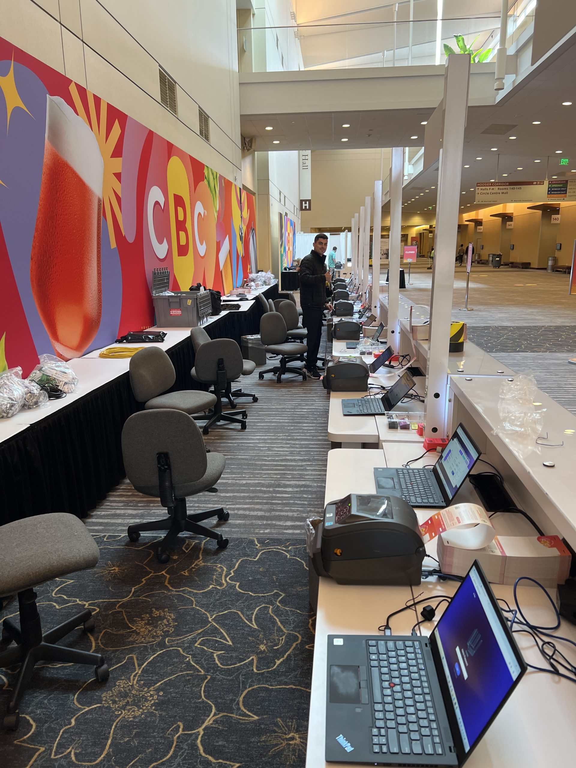 Row of check-in laptops and label printers in a convention hall with a CBC mural
