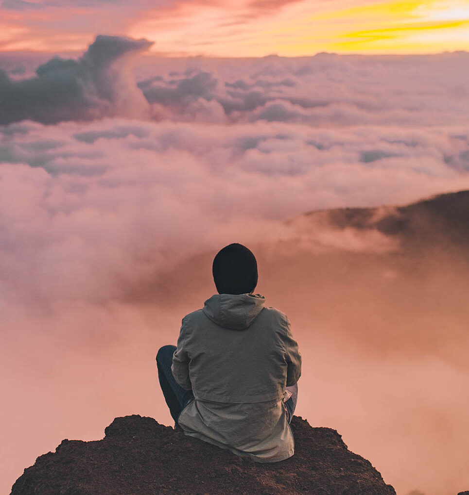 A person seated on a rocky peak looking out over a sea of clouds at a pink-orange sunset