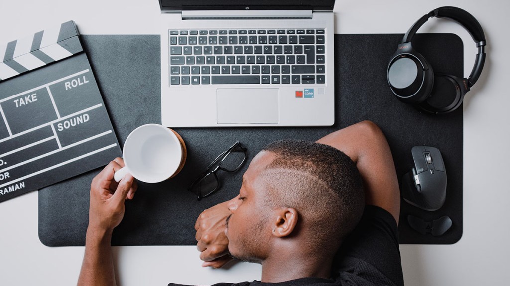 Overhead view of a man resting his head on a desk beside a laptop — burnout