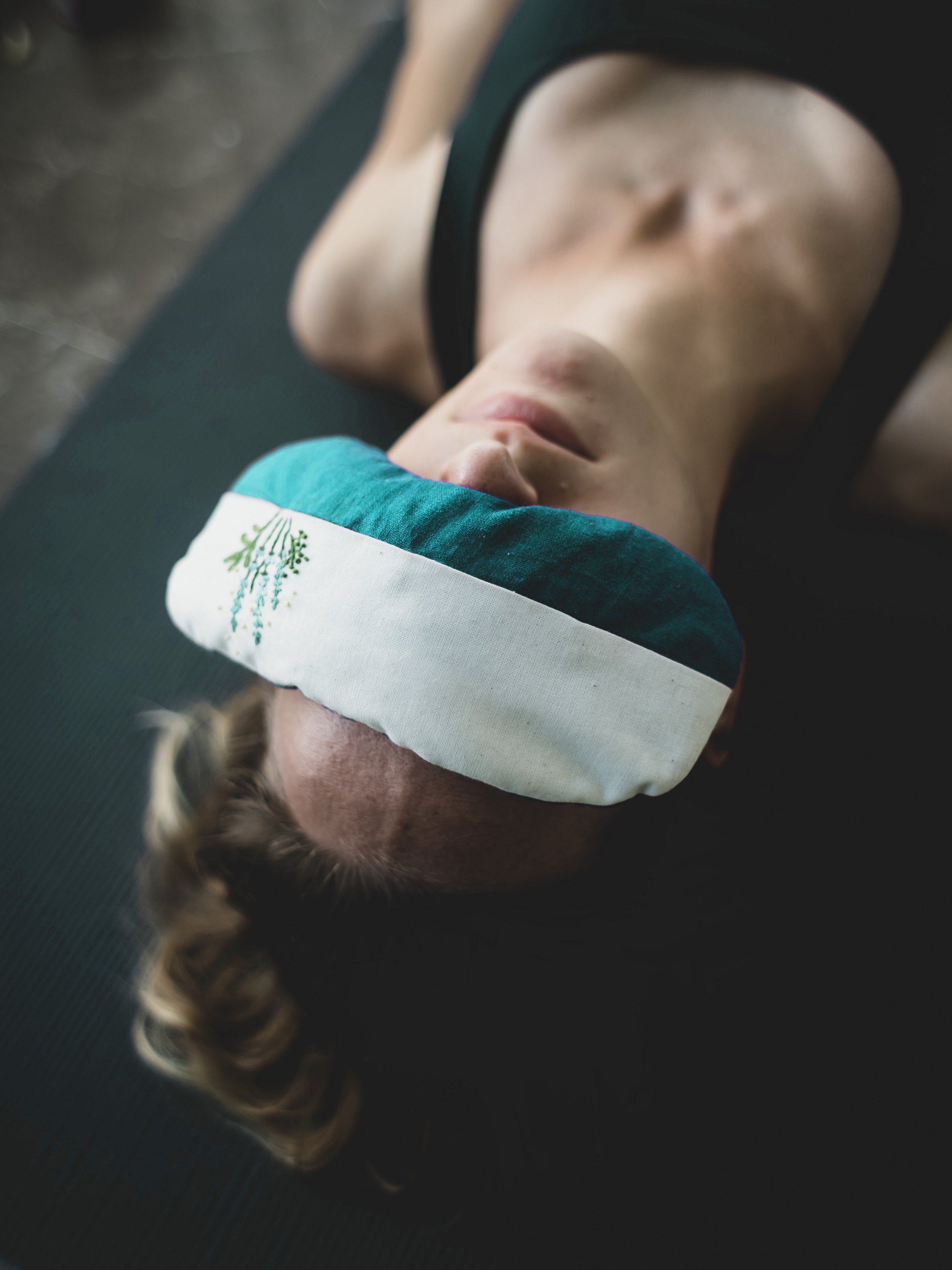 A woman lying on a yoga mat with a teal eye pillow covering her eyes