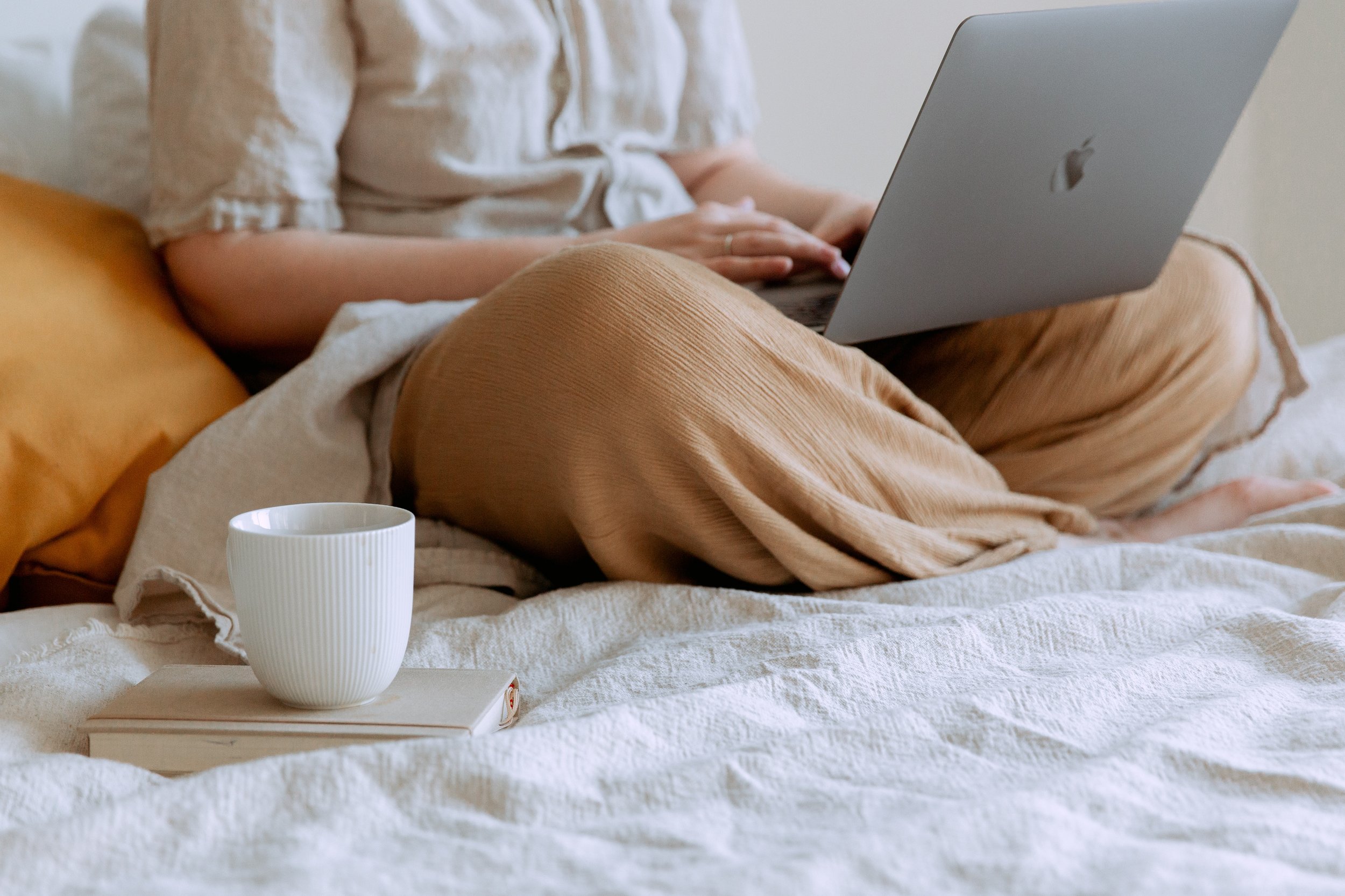 A person sitting cross-legged on a bed using a laptop, with a mug and book nearby