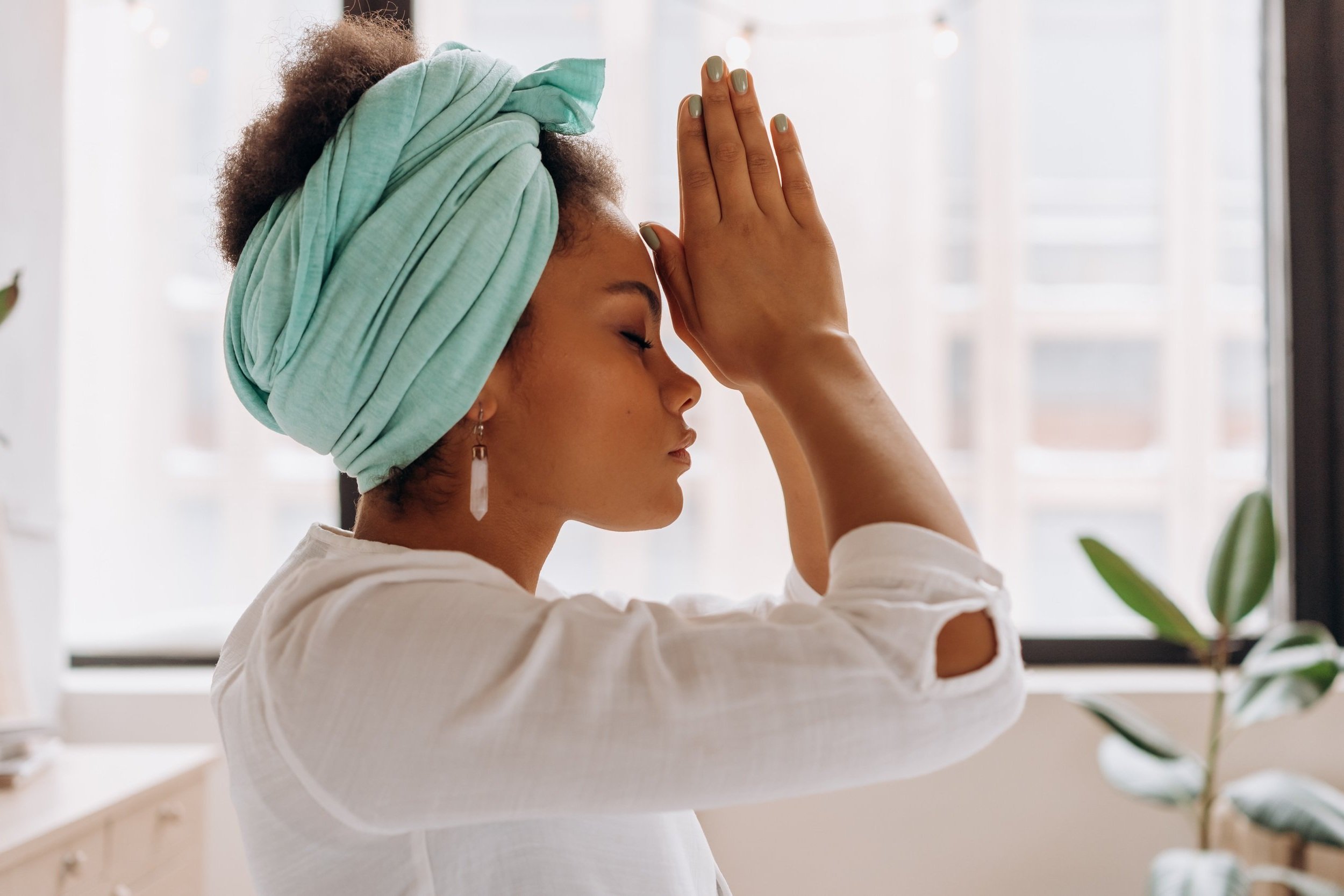 A woman in a teal head wrap with hands pressed together at her forehead in a mindful, centering pose