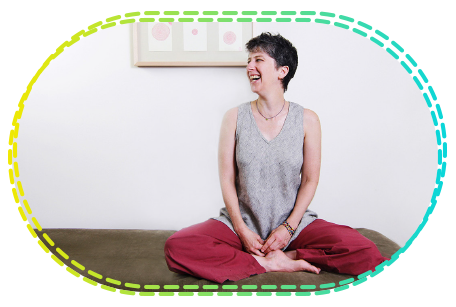 Clare Maxwell sitting cross-legged on a bench, laughing, in a bright room