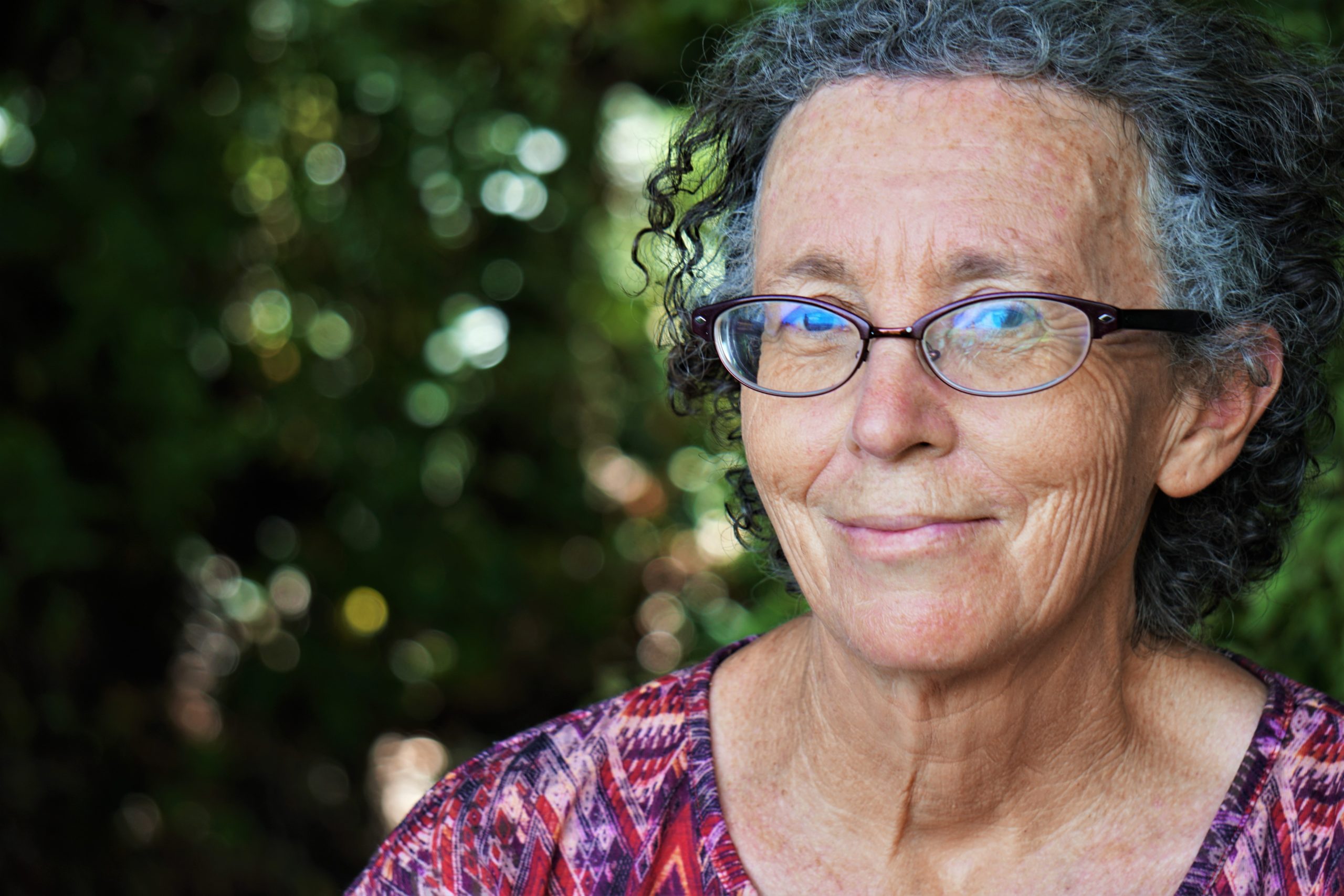 Smiling older woman with curly grey hair and glasses standing outdoors among green foliage