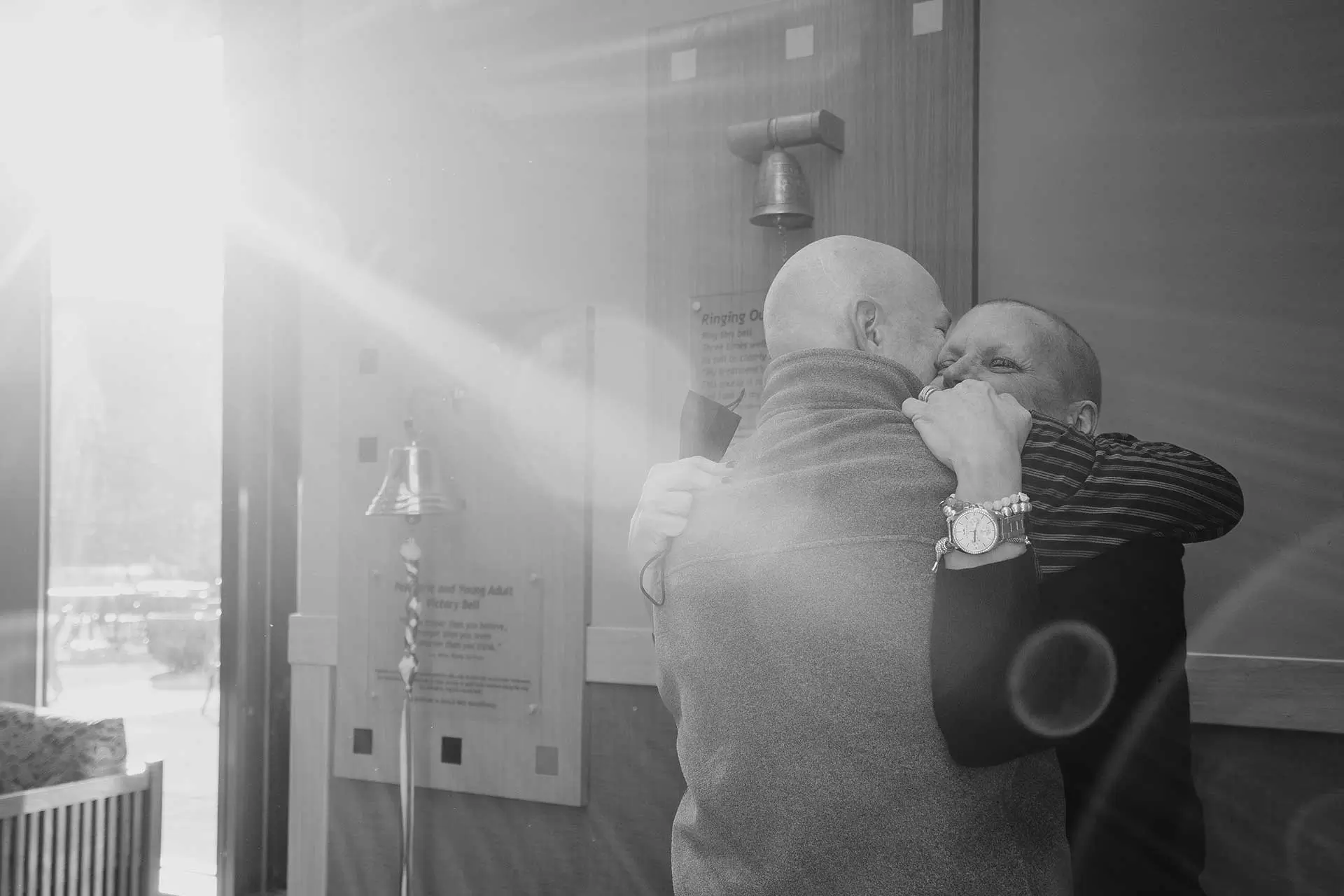 Two bald people embracing beside a cancer treatment victory bell