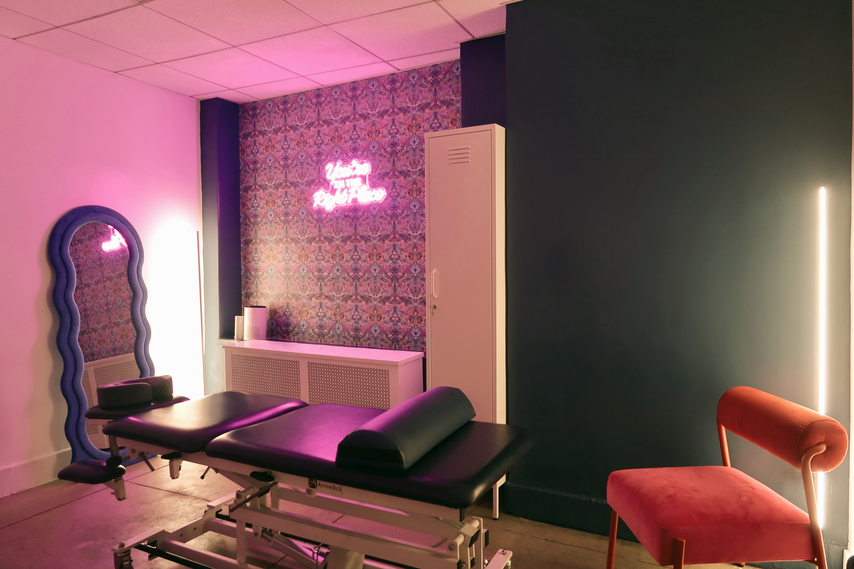 Encore treatment room with a purple neon sign, patterned wallpaper, treatment table and wavy mirror