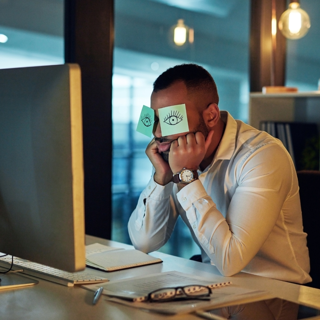 Exhausted manager at his desk with sticky-note eyes drawn over his face