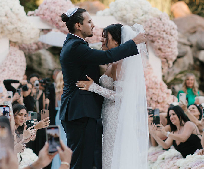 Outdoor wedding ceremony with a groom lifting a veil amid pink florals and guests