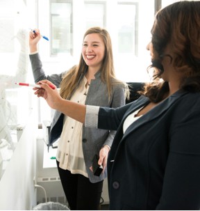 Two women writing on a whiteboard in an office, smiling
