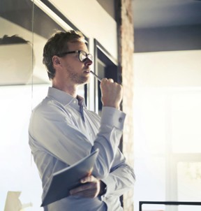 Leader in glasses holding a notebook, looking thoughtfully at a glass wall