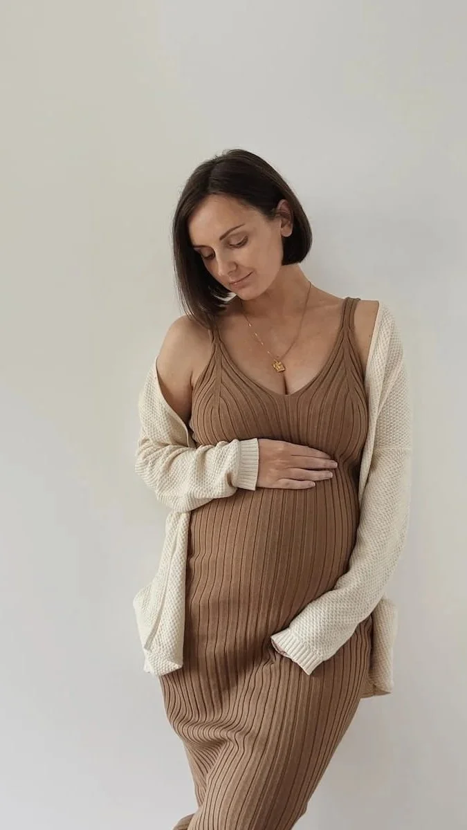 Pregnant woman in a brown ribbed dress and cream cardigan, cradling her bump.