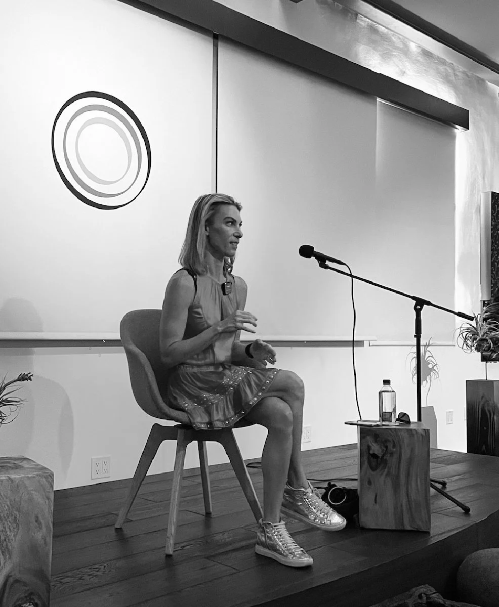 Debra Strougo seated on stage with a microphone, mid-talk, in black and white