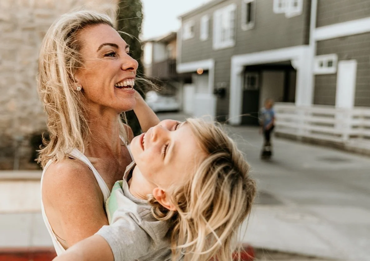 Debra Strougo laughing while holding her child outdoors on a sunlit street