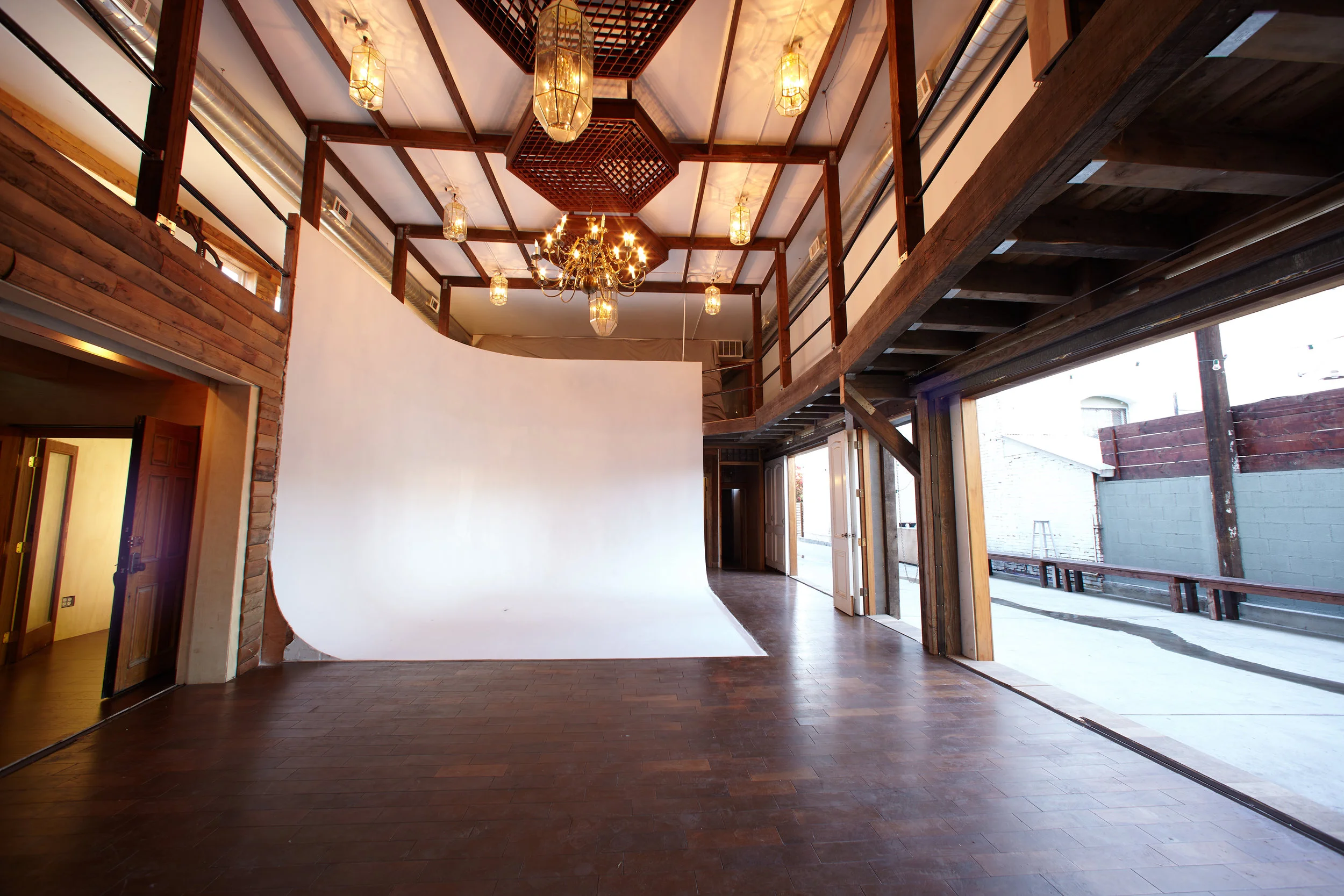 Studio East — open space with a white cyclorama backdrop, dark parquet floors, an exposed-beam mezzanine, and chandelier lighting