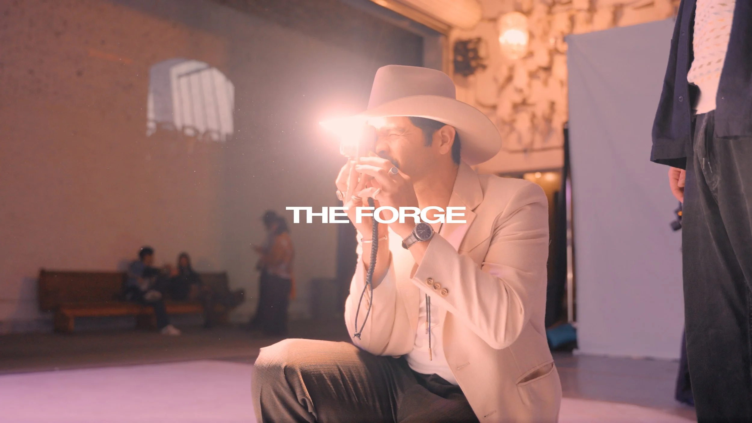 A person in a white cowboy hat and cream blazer crouching with a camera flash in the studio, with 'THE FORGE' overlaid