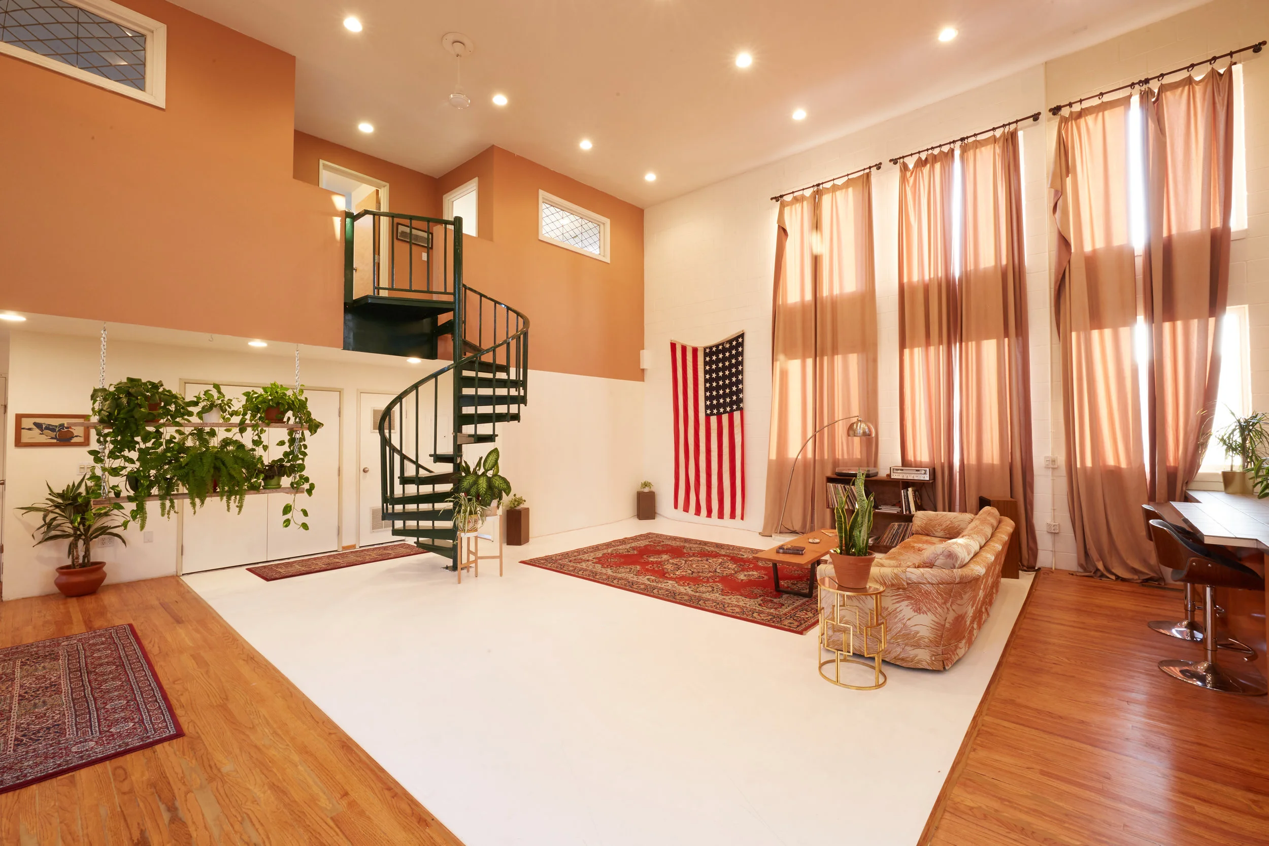 Loft West — warm terracotta walls, a spiral staircase, hanging plants, floor-length curtains, and a white backdrop area