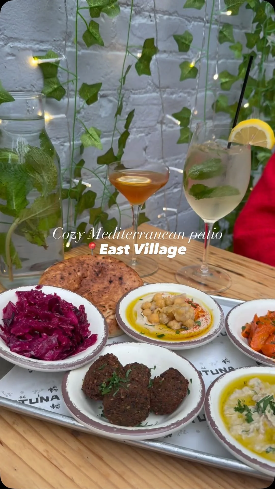 A tray of Mediterranean dishes including falafel, hummus, and salads with Aperol and Hugo spritzes on a wooden table on the patio.