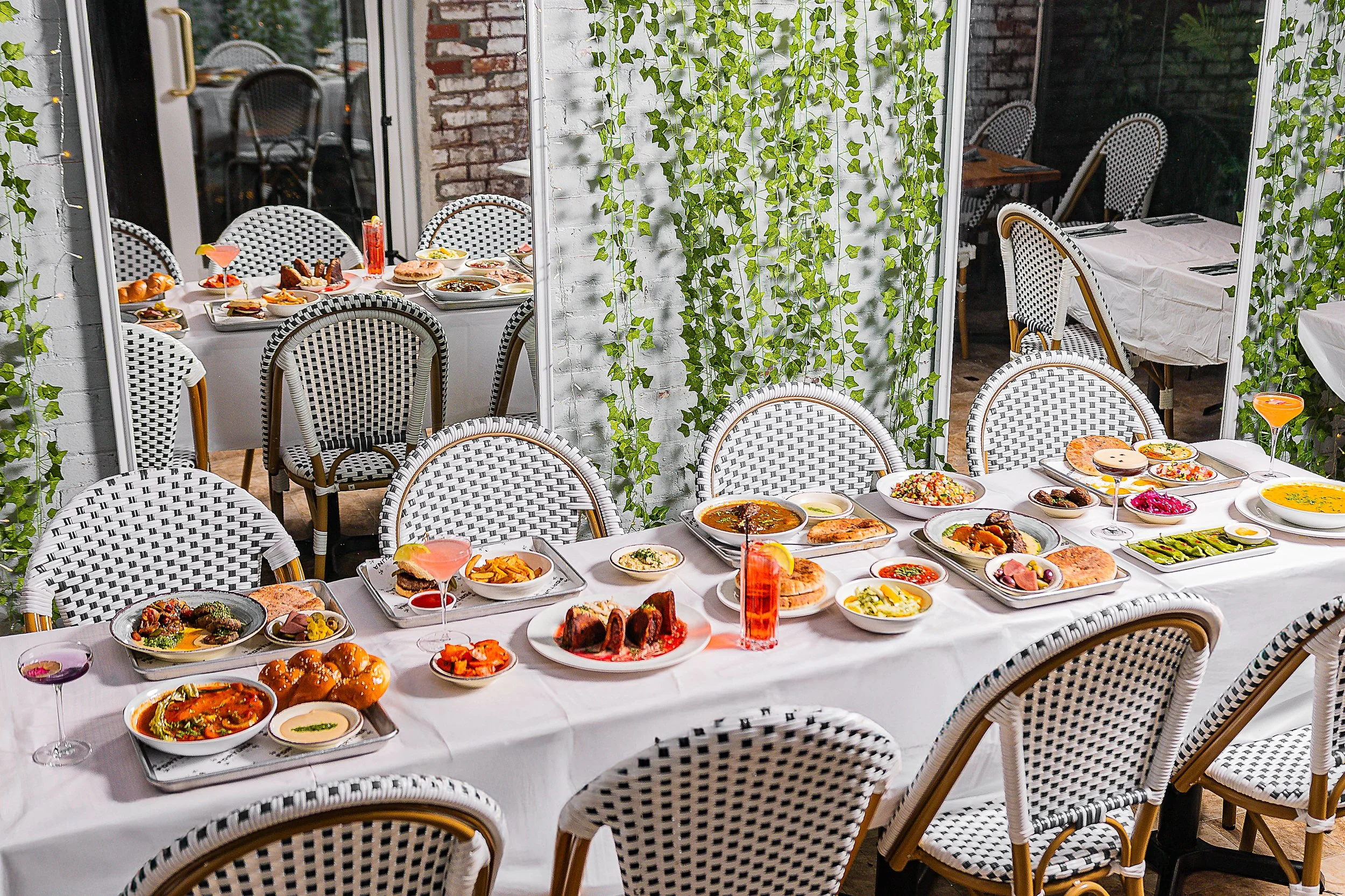 A dining table set with various dishes and drinks inside a bright room with large windows and green ivy hanging decor.