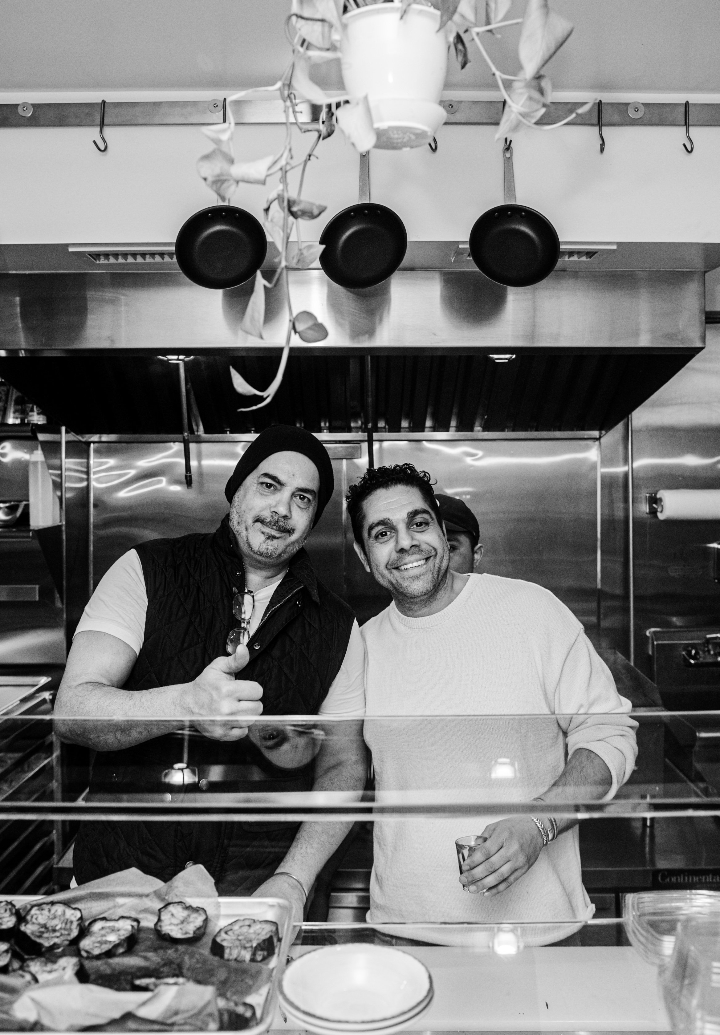 Two men standing behind a glass counter at Fortuna, smiling, with kitchen equipment behind them.
