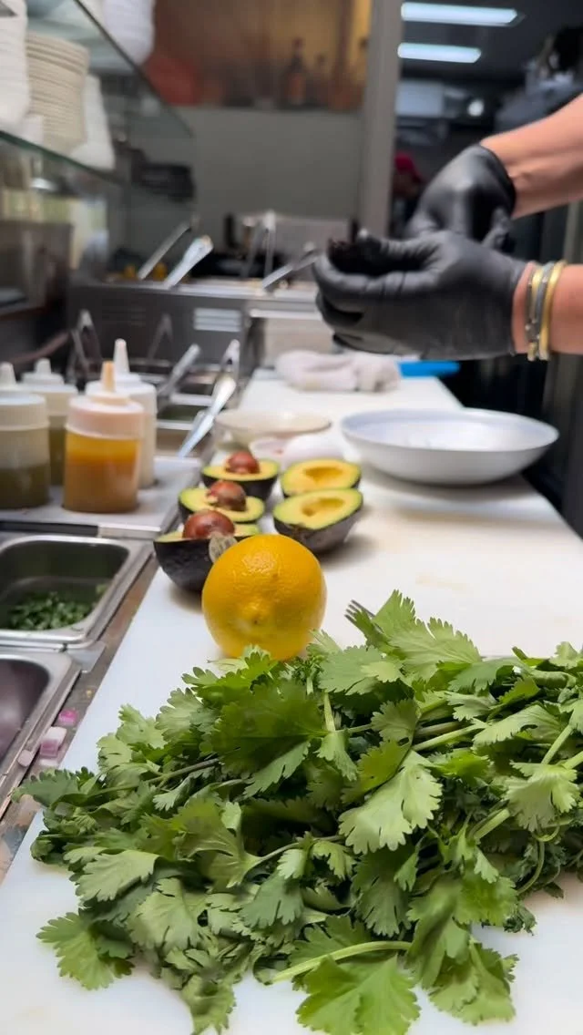Fresh cilantro, a lemon, and cut avocados on a kitchen counter with a person preparing food in the background.