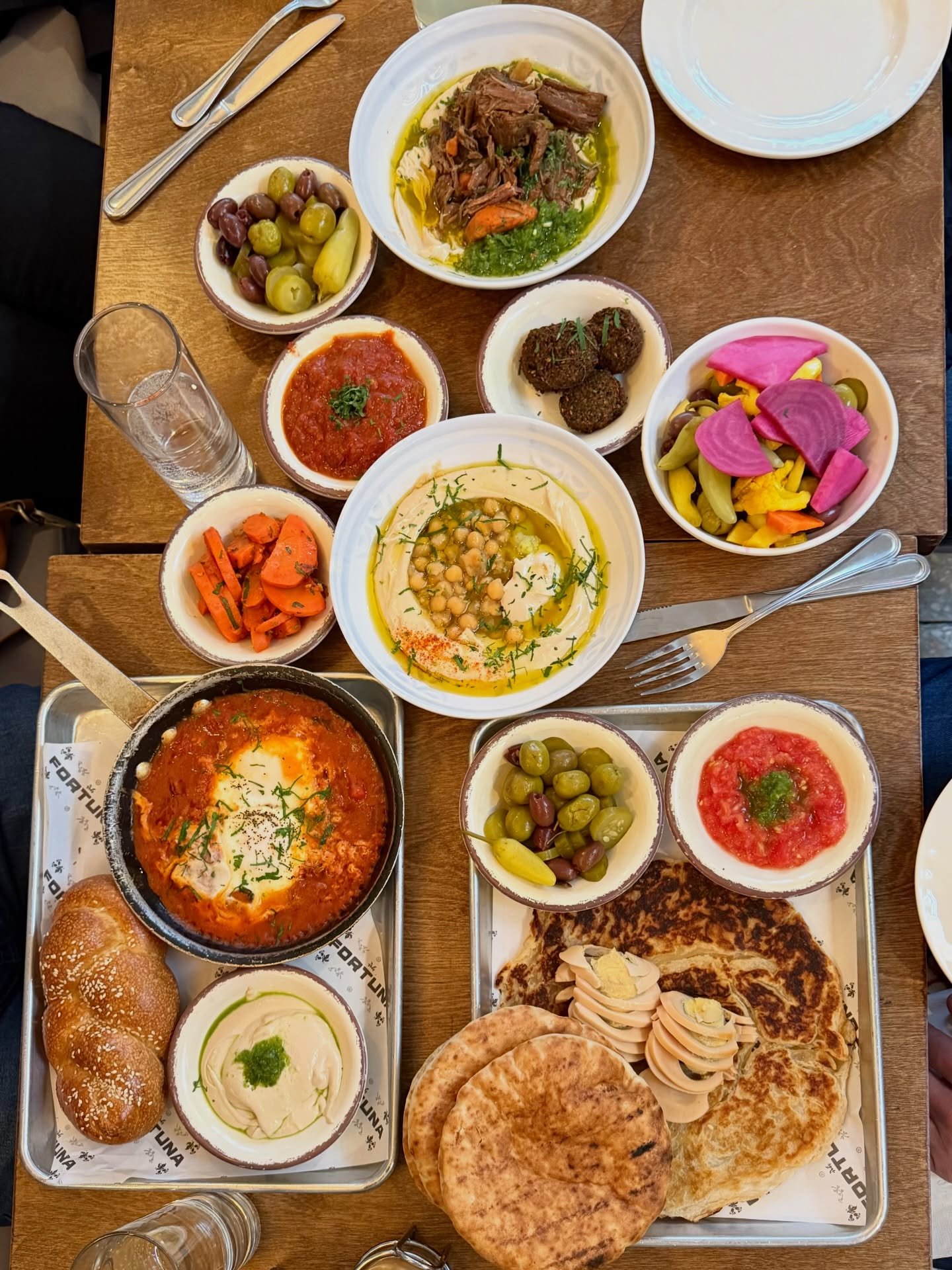 Assorted Middle Eastern dishes on a wooden table, including bread, hummus, tomato salad, olives, pickled vegetables and braised beef.
