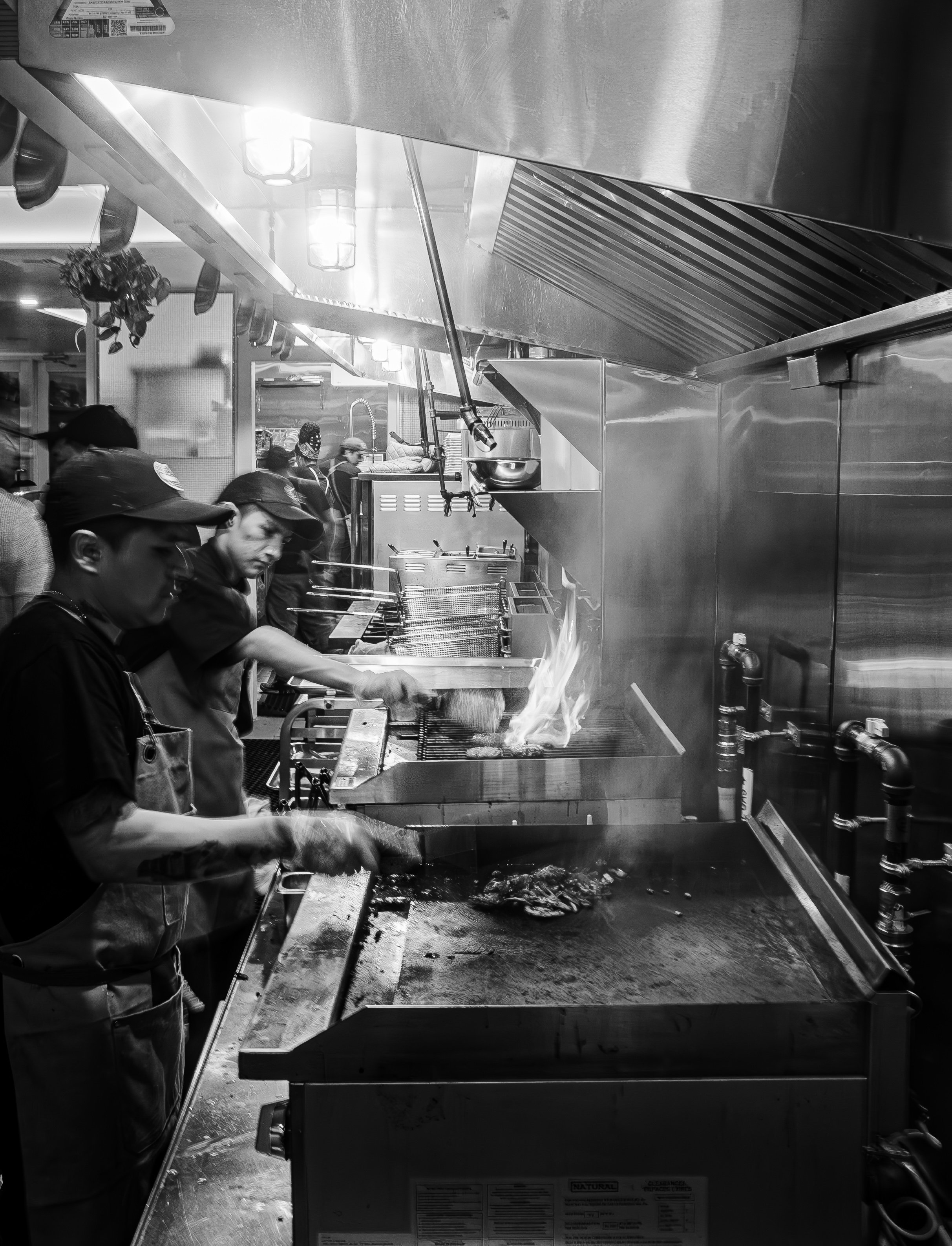 Kitchen scene with staff cooking; stovetop with flames, stainless steel surfaces, and multiple chefs.