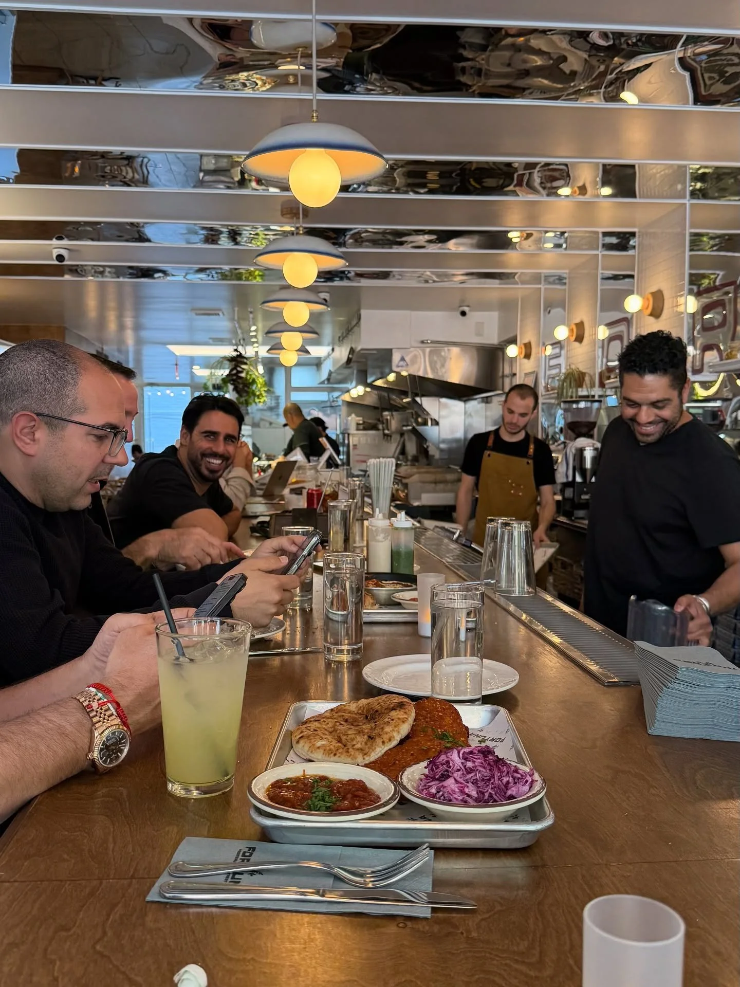 A group of four people dining at a restaurant counter, with plates of food and a busy kitchen in the background.
