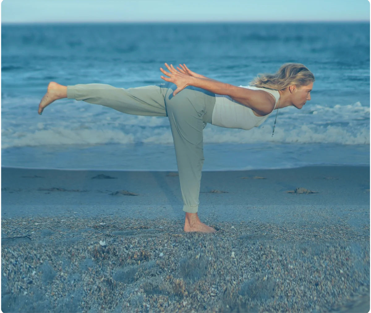 Woman performing a warrior III yoga balance pose on a beach shoreline