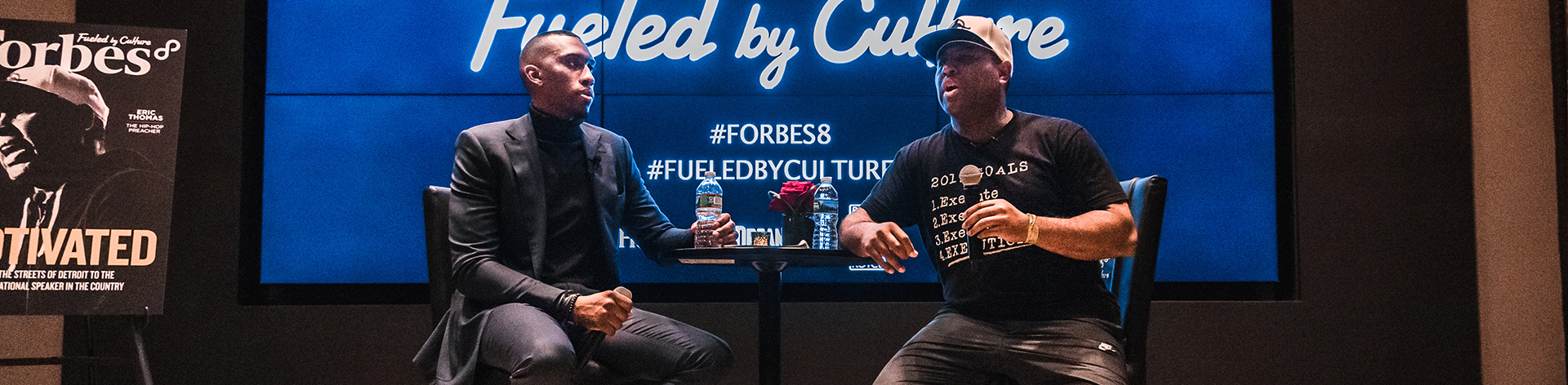 Two men on stage at a Fueled by Culture event in front of a screen with Forbes hashtags