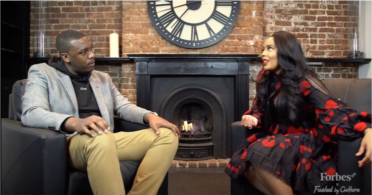 Two people seated by a fireplace in a brick-walled room in conversation, Forbes x Fueled by Culture branding