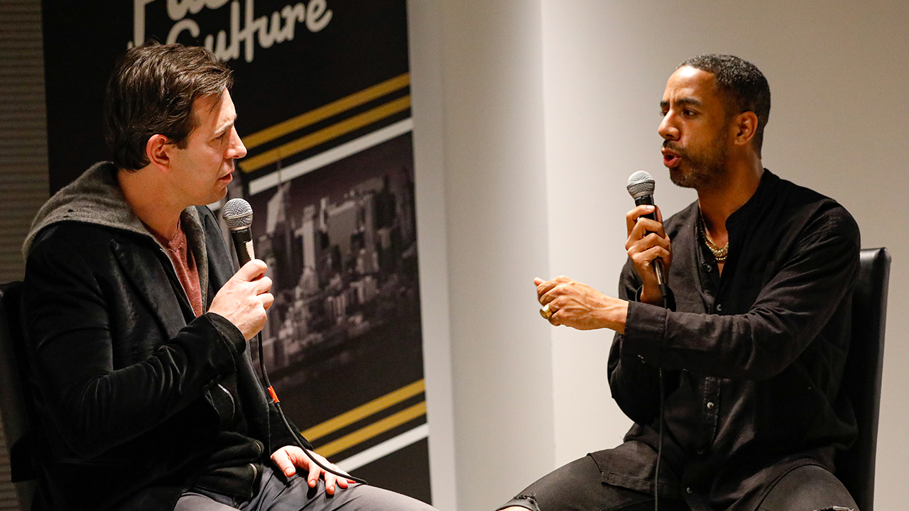 Two men seated facing each other holding microphones in front of a Fueled by Culture banner