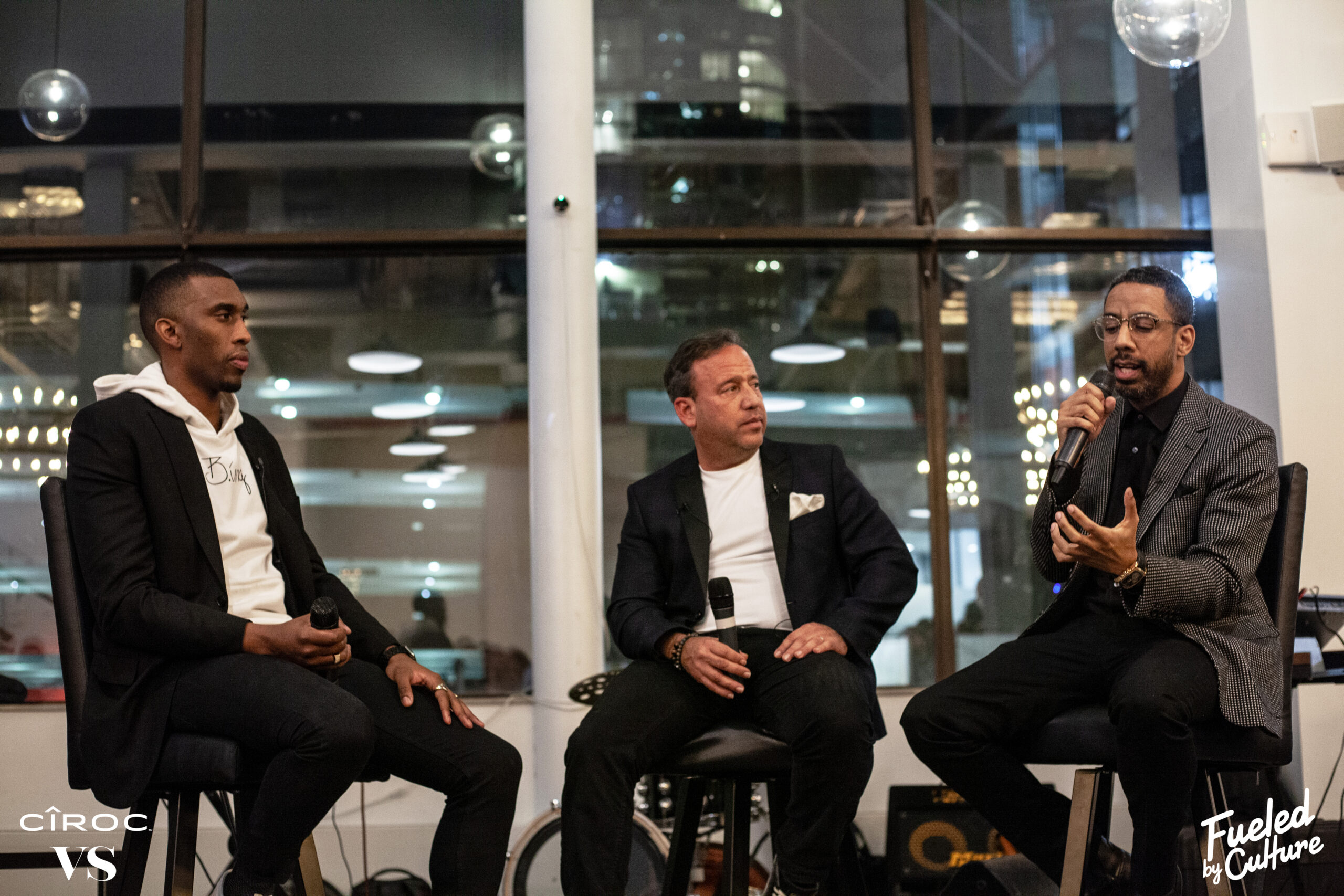 Three men seated on stools at a panel event with microphones and Fueled by Culture branding