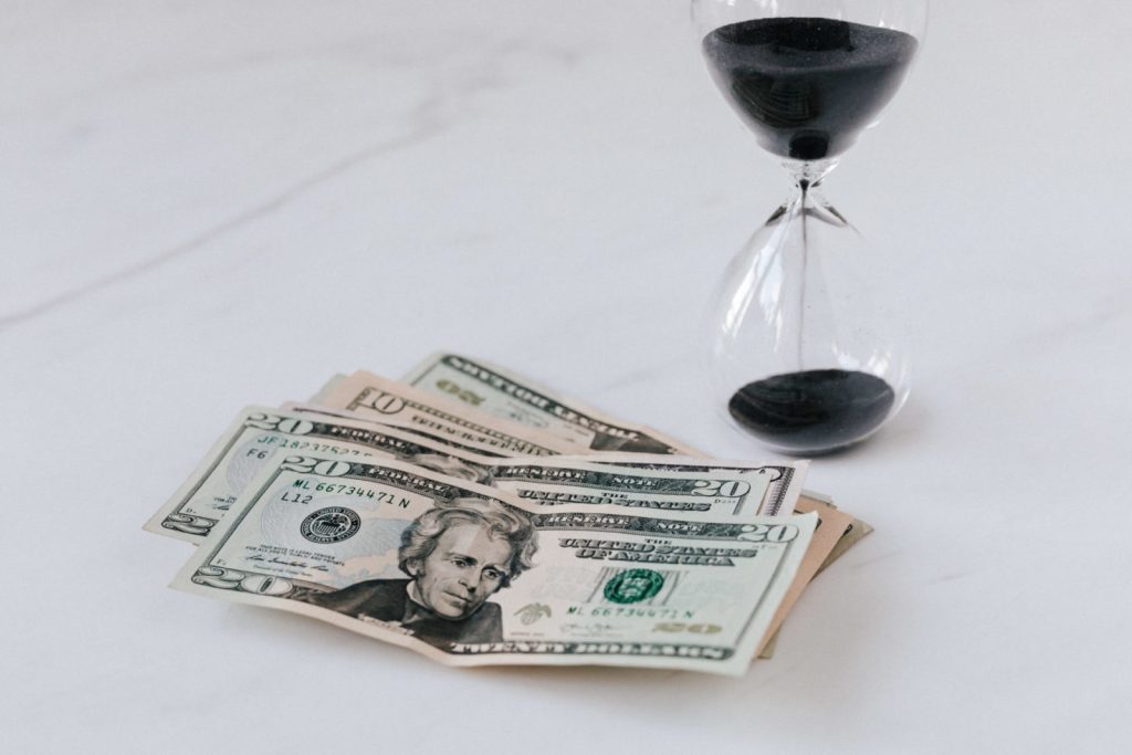 US dollar bills fanned out beside a black-sand hourglass on white marble — time is money