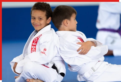 Two young boys in white gis sitting back to back on a blue mat, smiling