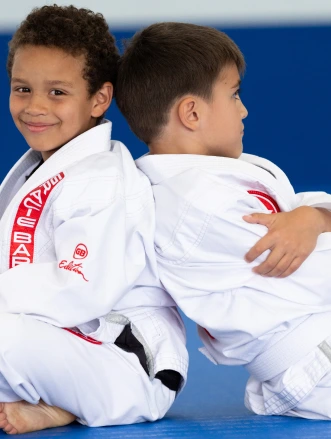 Two boys in white gis sitting back to back on a blue mat, smiling