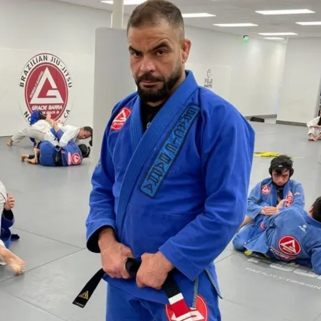 Fabio Trindade in a blue gi with black belt on the mat with kids training behind him