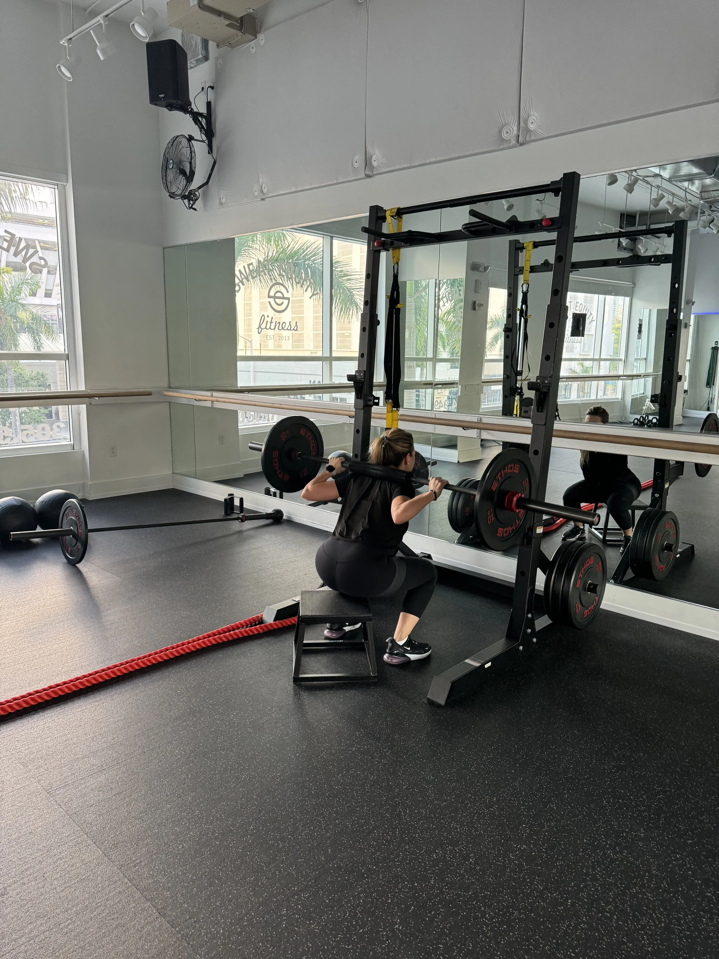 Woman performing a barbell squat on a power rack, Sweat Equity Fitness logo visible in the mirror