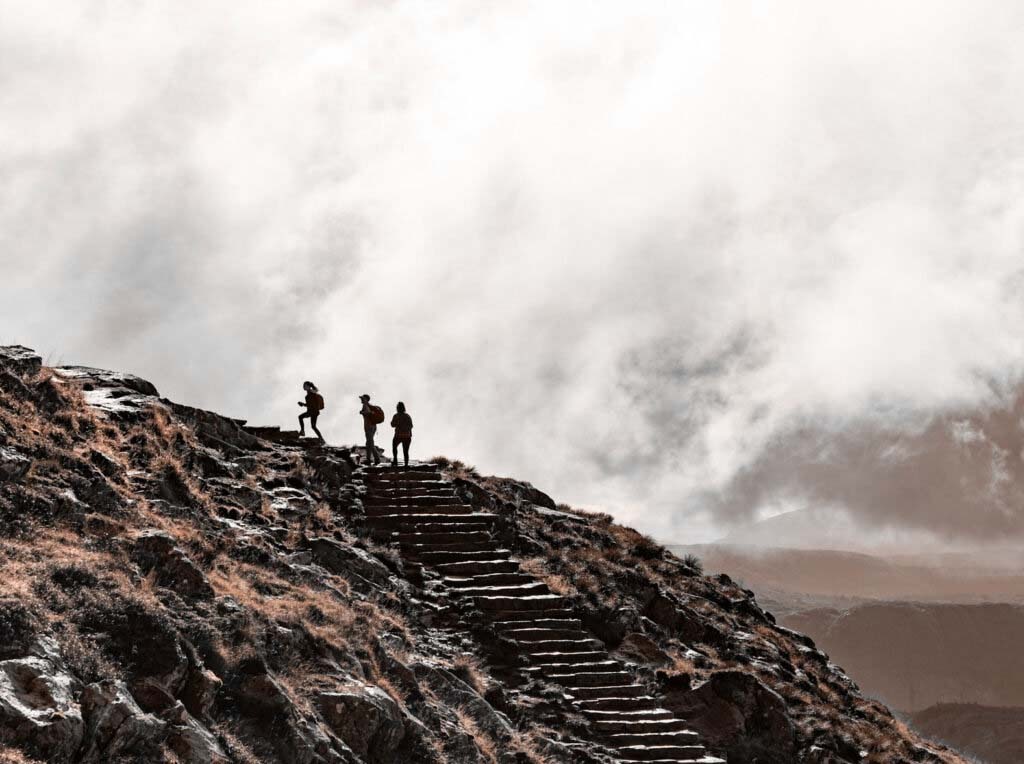 Three hikers silhouetted on stone steps climbing a rocky mountain peak into dramatic clouds